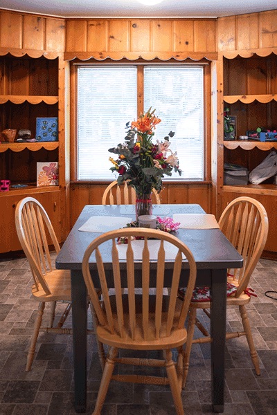A wooden dining table with four chairs and a flower vase, set in a cozy wood-paneled room with shelves.